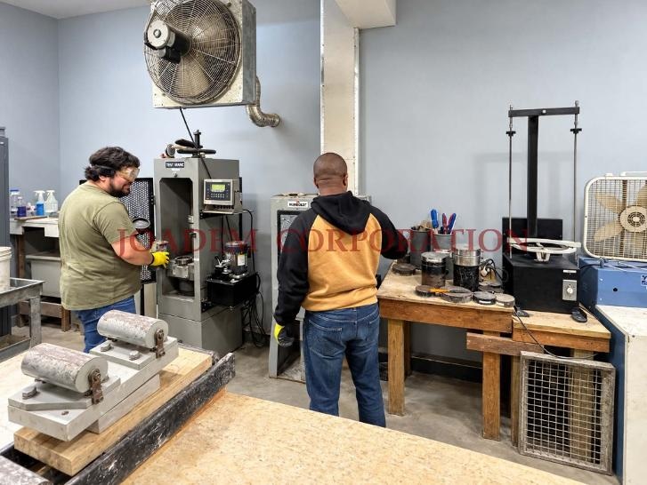 Two JOADEM technicians at a materials testing bench beside a compression machine, cylinder molds, and lab equipment